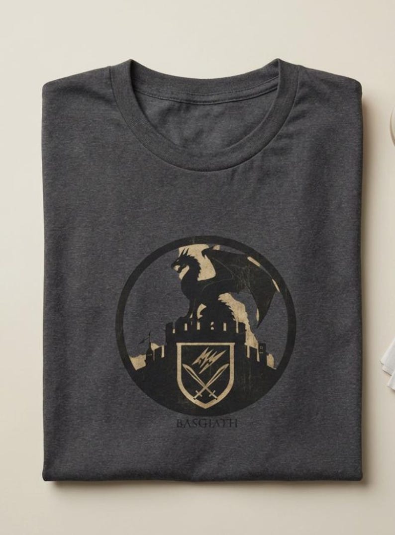 Puede incluir: Camiseta gris oscuro con un dise&ntilde;o circular. El dise&ntilde;o presenta un drag&oacute;n sobre un castillo, un escudo con espadas cruzadas y la palabra "BASGIATH" debajo. El dise&ntilde;o es en negro y beige.