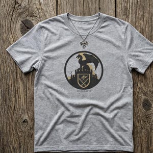 Puede incluir: Una camiseta gris con un dise&ntilde;o circular que representa un drag&oacute;n negro sobre un castillo, con la palabra "BASGIATH" debajo. Tambi&eacute;n se ve un collar plateado con un colgante de drag&oacute;n.