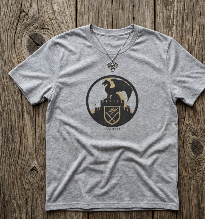 Puede incluir: Una camiseta gris con un dise&ntilde;o circular que representa un drag&oacute;n negro sobre un castillo, con la palabra "BASGIATH" debajo. Tambi&eacute;n se ve un collar plateado con un colgante de drag&oacute;n.