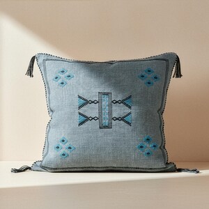 May include: A square, pale blue cushion with a geometric embroidered design in black and turquoise. The cushion has black tassels on each corner and a black stitched border. The cushion is made of a textured fabric.