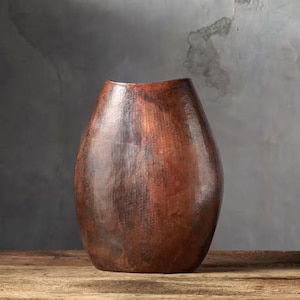 Op de afbeelding: Een grote, koperkleurige vaas met een brede, afgeronde body en een licht uitlopende opening. Het oppervlak heeft een getextureerd, gehamerd uiterlijk, met kleurvariaties van licht- tot donkerbruin. De vaas is een decoratief item.