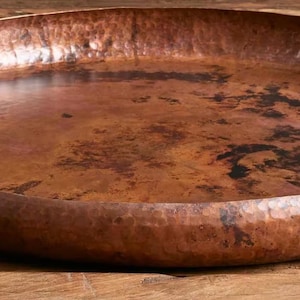 Pode incluir: Uma grande bandeja de servir de cobre martelado com uma borda larga e arredondada. A bandeja tem uma cor marrom quente e rica com &aacute;reas mais escuras, criando uma apar&ecirc;ncia r&uacute;stica e texturizada. A bandeja est&aacute; em uma superf&iacute;cie de madeira.