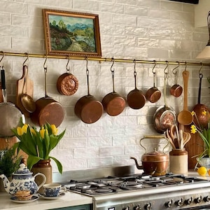 May include: A white tiled kitchen with a copper pot rack hanging above a gas hob. The rack is filled with copper pots and pans of various sizes. There is a framed picture hanging on the wall above the rack. A bouquet of yellow tulips sits on the counter in front of the hob.