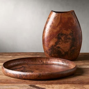Pode incluir: Um vaso decorativo em tons de cobre e uma bandeja redonda est&atilde;o sobre uma superf&iacute;cie de madeira. O vaso tem uma forma alongada &uacute;nica com uma abertura larga. A bandeja &eacute; rasa e tem uma textura martelada, refletindo a luz. Os itens t&ecirc;m uma apar&ecirc;ncia r&uacute;stica e artesanal.