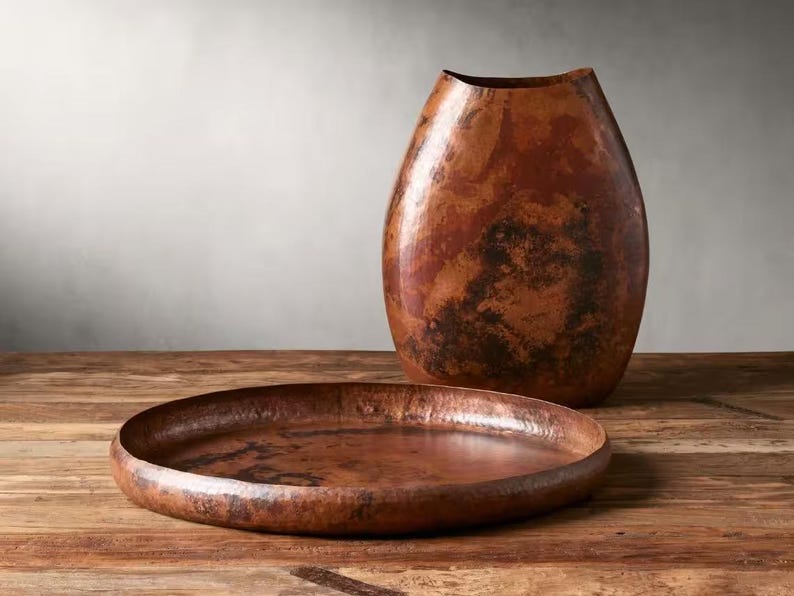 Pode incluir: Um vaso decorativo em tons de cobre e uma bandeja redonda est&atilde;o sobre uma superf&iacute;cie de madeira. O vaso tem uma forma alongada &uacute;nica com uma abertura larga. A bandeja &eacute; rasa e tem uma textura martelada, refletindo a luz. Os itens t&ecirc;m uma apar&ecirc;ncia r&uacute;stica e artesanal.