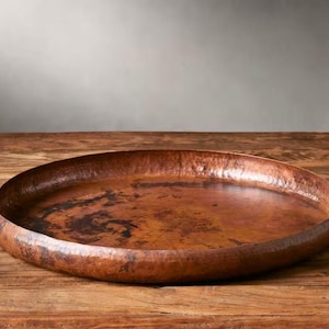 Może przedstawiać: Duża, młotkowana miedziana taca o szerokim, płytkim kształcie. Taca ma bogaty, czerwonobrązowy kolor i stoi na drewnianej powierzchni. Powierzchnia tacy wykazuje zmienność koloru i faktury.