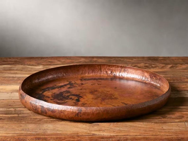 Pode incluir: Uma grande bandeja de cobre martelado com um design amplo e raso. A bandeja tem uma rica cor marrom avermelhada e assenta sobre uma superf&iacute;cie de madeira. A superf&iacute;cie da bandeja mostra varia&ccedil;&otilde;es de cor e textura.