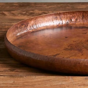 Pode incluir: Uma grande bandeja de servir em cobre martelado com formato oval raso. A bandeja tem uma cor marrom quente e rica e uma superf&iacute;cie texturizada. A borda &eacute; ligeiramente elevada e arredondada. A bandeja &eacute; exibida em uma superf&iacute;cie de madeira.