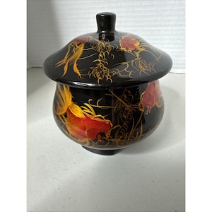 May include: A black, round decorative jar with a lid. The jar and lid are adorned with gold and red floral designs. The jar has a small, black knob on top.