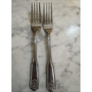 May include: Two silver-toned metal forks featuring four tines each. The handles have a vertical ribbed design and a decorative shell-like motif at the base. The forks have a polished finish and a slender neck connecting the tines to the handle.