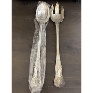 May include: A silver serving spoon and fork set. The spoon is in a plastic sleeve. Both utensils have ornate, floral-like designs on the handles. The fork has three tines. The set is on a dark brown wooden surface.