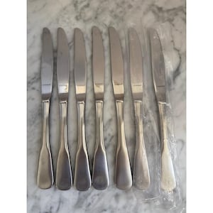 May include: A set of seven silver-toned stainless steel dinner knives arranged in a row. Each knife features a smooth, rounded handle and a serrated blade with the brand name Oneida stamped clearly on the metal surface of the blade.