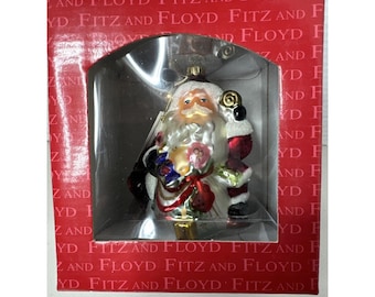 May include: A Fitz and Floyd Christmas ornament featuring Santa Claus in a red and white suit, holding a small gift and a staff. The ornament is inside a red and white box with the words "Fitz and Floyd" printed on it.