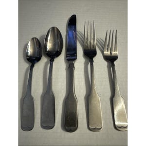 May include: A set of five silver-colored utensils: two spoons, a knife, and two forks. The spoons have rounded bowls, the knife has a curved blade, and the forks have four tines. The utensils are arranged vertically on a light-colored surface.