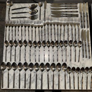May include: A complete set of vintage silverware, including spoons, forks, knives, and a ladle, arranged on a marble surface. The silverware has a decorative pattern on the handles and a polished finish, ideal for formal dining.