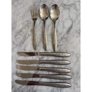 May include: A set of stainless steel flatware consisting of one fork, two spoons, and five knives. Each piece features a polished metallic finish and a decorative floral pattern embossed on the handle, providing a consistent design across the set.