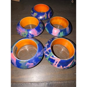May include: A set of five circular napkin rings featuring a vibrant blue exterior with abstract pink and green paint splatters. Each ring has a smooth, bright orange interior lining and a rounded, thick profile for holding napkins securely.