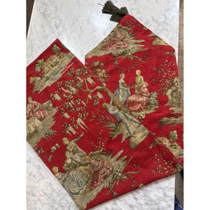 May include: A red table runner featuring a woven pastoral scene with figures in period clothing, trees, and buildings in shades of gold, green, and blue. The runner is finished with a dark green tassel at the pointed end, showcasing a detailed tapestry-style fabric texture throughout the entire piece.