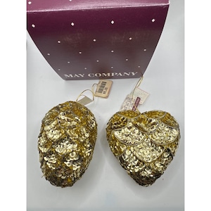 May include: Two gold sequined ornaments, one shaped like a pinecone and one shaped like a heart, each with a gold hanging string and a price tag. They are positioned in front of a dark purple box with white diamond patterns and the text MAY COMPANY.