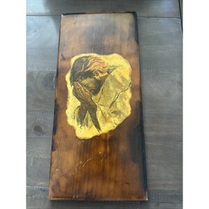 May include: A rectangular wooden plaque with a distressed finish. The plaque features a central image of a person with their face in their hands, rendered in shades of yellow, gold, and brown. The artwork is set against a dark wood background.