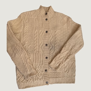 May include: A beige long-sleeve cardigan featuring a complex cable knit pattern throughout the body and sleeves. The garment has a high collar and a front button closure with six dark circular buttons aligned vertically down the center.