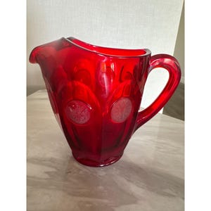 May include: A vibrant red glass pitcher featuring a series of embossed circular medallions around the exterior body. The pitcher has a wide pouring spout, a curved handle on the right side, and a translucent finish that catches the light.