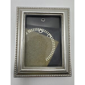 May include: A rectangular silver-toned picture frame with a beaded border design. The frame features a clear glass front covering a dark interior display area that contains a textured, light beige foam insert with several small circular metal eyelets.