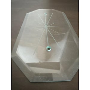 May include: A clear, octagonal glass plate featuring a central etched starburst design. The glass has beveled edges and two circular holes positioned along the vertical axis, with one hole featuring a small metal ring insert for mounting.