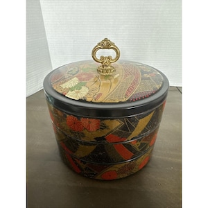 May include: A three-tiered, round, black and gold decorative container with a clear lid. The container is adorned with floral and geometric patterns in red, gold, and white. It features a gold handle on top.