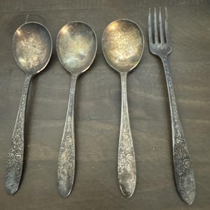May include: Four antique silver-toned utensils: three spoons and one fork. The spoons have oval bowls and floral patterned handles. The fork has four tines and a matching floral design. The silverware is arranged on a dark wooden surface.