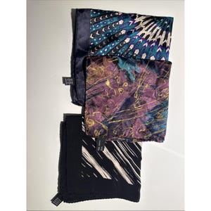 May include: Three folded silk scarves in a stack. The top scarf has a teal and purple feather design. The middle scarf is purple with gold swirls. The bottom scarf is black with white diagonal lines. Each scarf has a small tag.