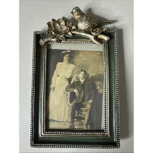 May include: A vintage picture frame with a black and white photograph of a couple. The frame is green with a silver beaded border and a decorative silver bird and flower on top. The photograph is of a man seated and a woman standing.