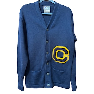 May include: Navy blue cardigan sweater with a V-neck, button closure, and two front pockets. Features a large yellow and navy blue letter "C" patch on the left side. The label reads "Theurer".