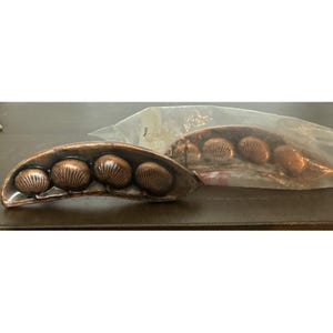May include: Two bronze-colored decorative drawer pulls, each shaped like a pea pod, with four seashell-shaped details. One is loose, and the other is in a clear plastic bag. The pulls are on a dark brown surface.