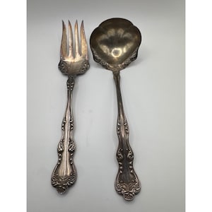 May include: A silver-toned metal serving fork and a matching serving spoon with a scalloped bowl. Both utensils feature ornate, embossed scrollwork patterns along the handles and the neck, with a tarnished finish throughout the metal surfaces.