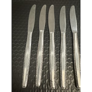 May include: Set of five silver-colored butter knives. Each knife features a decorative, ornate pattern along the handle's edge. The blades are smooth and curved. The knives are arranged on a dark, textured surface.