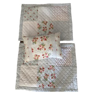 May include: A quilted pillow and two placemats with a floral pattern. The placemats and pillow have a light blue background with patches of white, pink, and purple floral designs. The placemats and pillow are quilted with a diamond pattern.