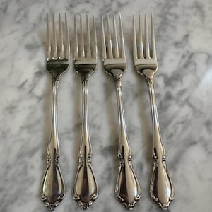May include: Four silver forks with ornate handles are arranged on a marble surface. The forks have a classic design with tines and decorative detailing on the handles. The silverware is likely for formal dining.