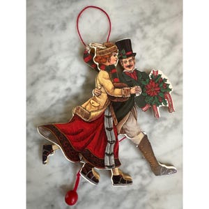 May include: A vintage Christmas ornament depicting a couple dancing. The woman wears a red skirt and tan coat, while the man wears a black top hat and holds a poinsettia. A red string is attached for hanging.