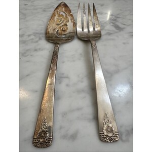 May include: A silver-toned metal cake server and a matching large fork. The cake server features a pointed, perforated head with a floral cutout. Both utensils have a tapered handle decorated with a raised floral motif at the base.