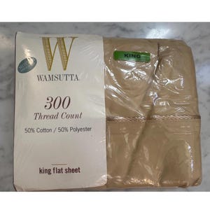 May include: A tan king flat sheet packaged in clear plastic. The label reads Wamsutta, 300 Thread Count, 50% Cotton / 50% Polyester, and Select Irregulars. The sheet features a decorative horizontal trim detail across the upper portion of the fabric.