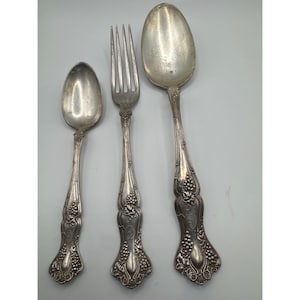 May include: A set of three silver-toned metal utensils consisting of a small spoon, a four-tined fork, and a large spoon. Each piece features an ornate embossed pattern of grape clusters and vine leaves along the handle and neck area.