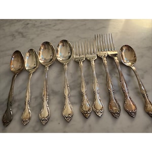 May include: A collection of antique silverware, including spoons and forks, arranged on a marble surface. The silverware has ornate, detailed handles and a polished, reflective finish, suggesting a vintage or heirloom quality.
