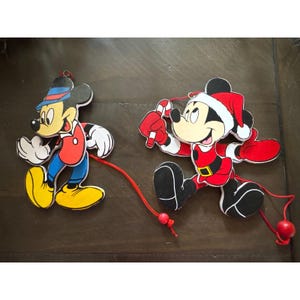 May include: Two flat wooden ornaments featuring Mickey Mouse. One figure wears a blue hat, red jacket, and blue pants. The second figure wears a Santa hat, red suit, and holds a candy cane. Both ornaments have red hanging strings with beads.