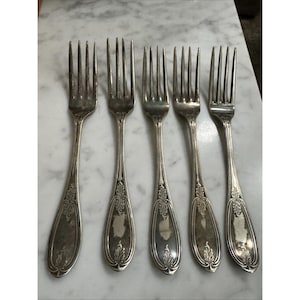 May include: Five antique silver forks arranged in a row. Each fork has a long, slender handle with an ornate floral design and four tines. The forks are displayed on a white marble surface, showcasing their vintage aesthetic.