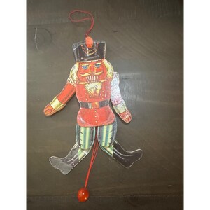 May include: A flat, articulated nutcracker ornament made of layered material, featuring a red jacket, black hat, and green trousers. The figure has movable limbs connected by strings, with a red pull cord and bead at the bottom for movement.