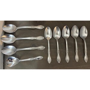 May include: A collection of silver spoons arranged on a dark surface. The spoons have ornate, detailed handles and rounded bowls. The spoons are likely for serving or eating, and the arrangement suggests a set.
