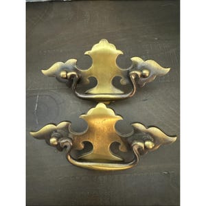 May include: Two antique brass drawer pulls with a decorative design. Each pull features a central handle and ornate, stylized cloud-like accents. The brass has a warm, aged patina, adding to the vintage aesthetic. These pulls are ideal for furniture restoration projects.