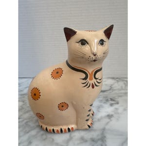 May include: A ceramic cat figurine with a cream-colored base, featuring hand-painted orange sunburst patterns on its body, black and orange striped accents on its paws and tail, and a decorative black and orange collar around its neck.