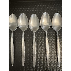 May include: Five silver spoons arranged in a row. Each spoon has a rounded bowl and a long handle with a decorative pattern. The spoons are made of a shiny, reflective metal, likely stainless steel.